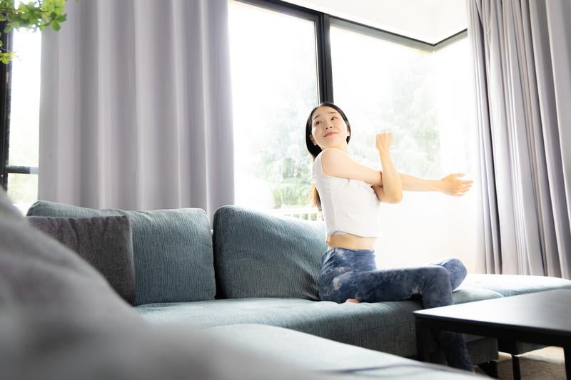 大きな窓のあるリビングでソファーの上でストレッチする女性の写真