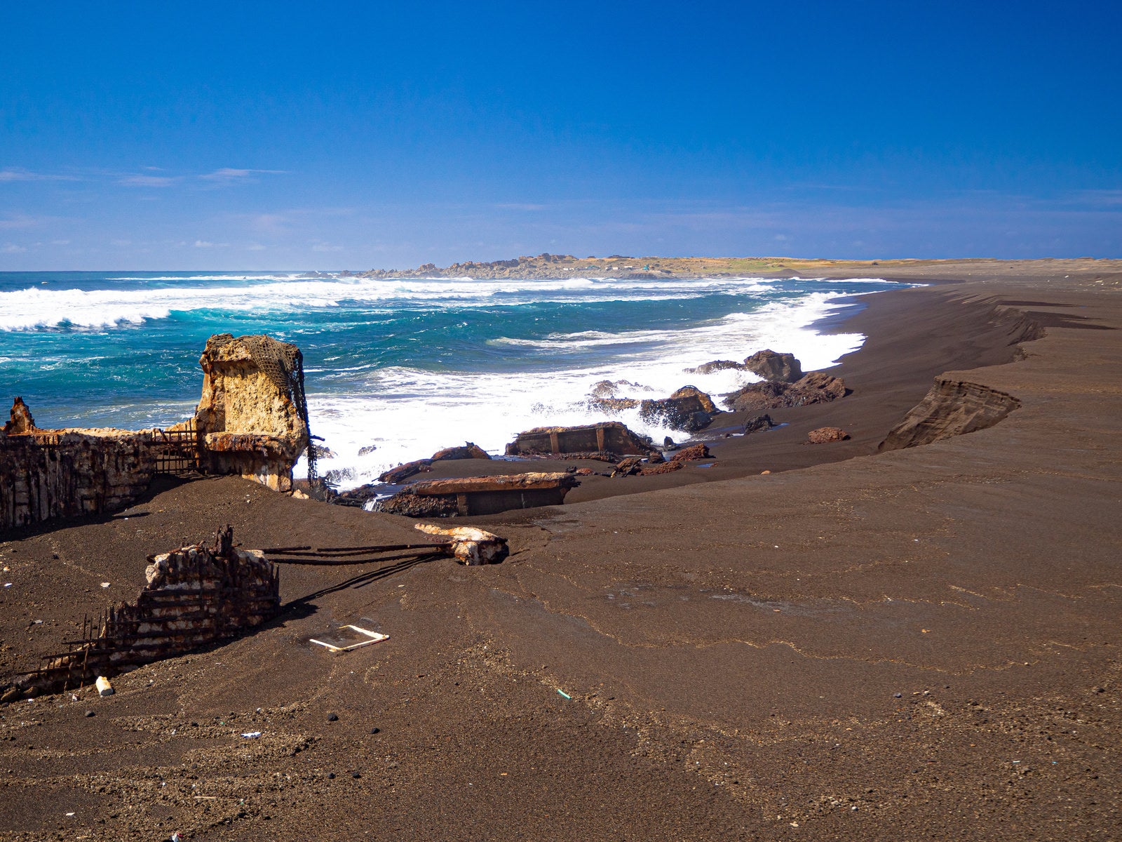 「千鳥ヶ浜の波打ち際で波に洗われる船の残骸と奥に見える釜岩」の写真