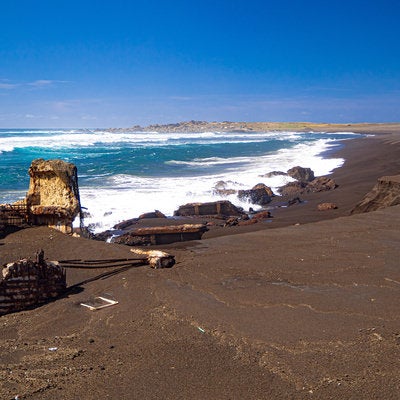 千鳥ヶ浜の波打ち際で波に洗われる船の残骸と奥に見える釜岩の写真