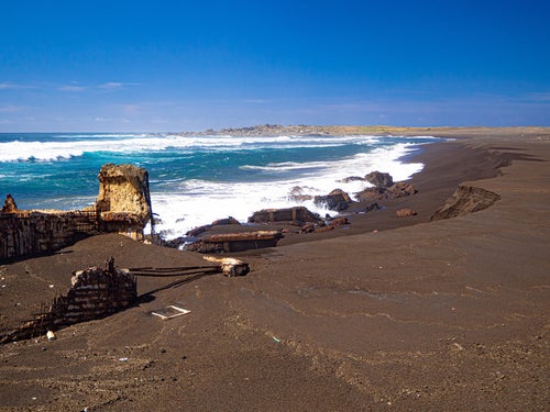 千鳥ヶ浜の波打ち際で波に洗われる船の残骸と奥に見える釜岩の写真