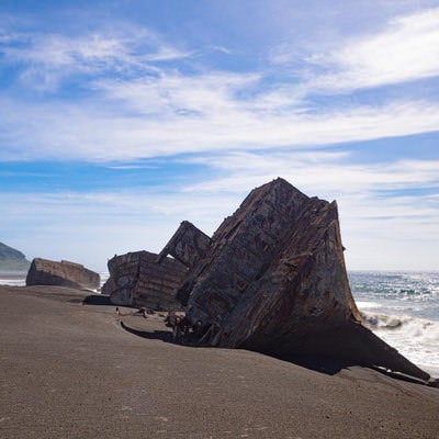 千鳥ヶ浜の波打ち際に横たわる朽ちたコンクリート船の瓦礫と遠くに見える摺鉢山の写真