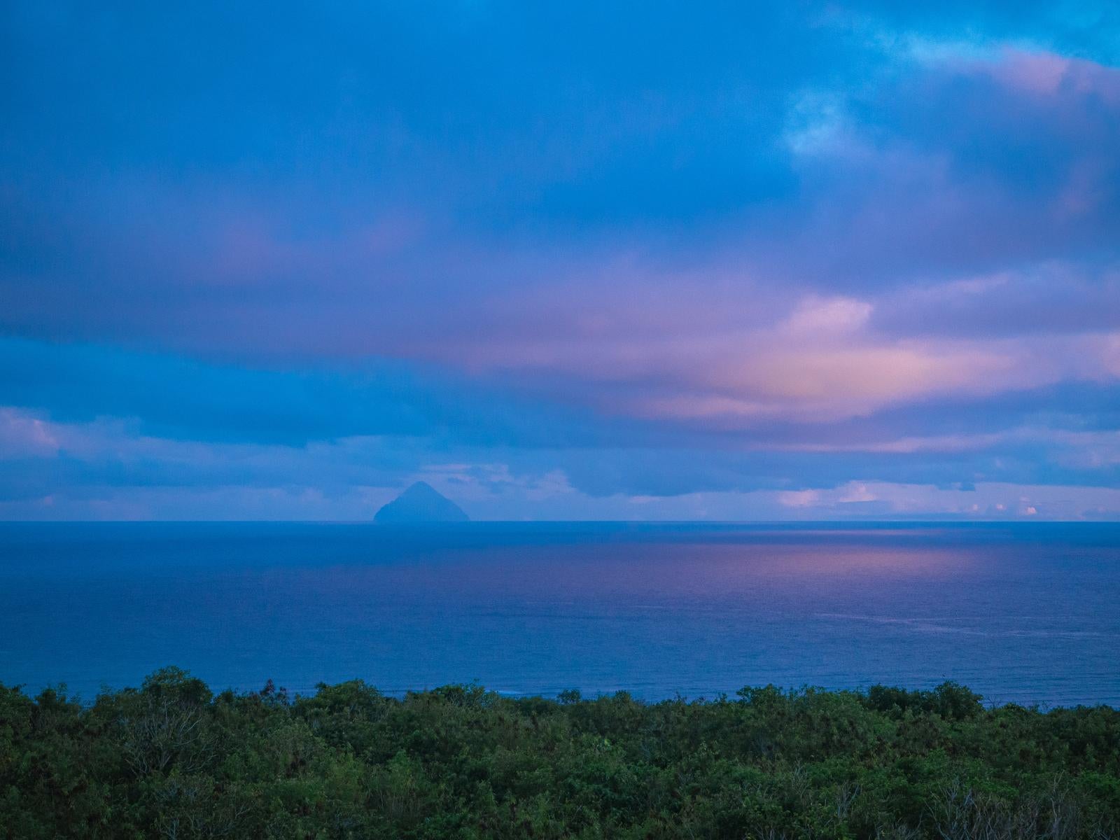 「夕焼け色の空と同じ色に染まる海の先にかすむ南硫黄島」の写真