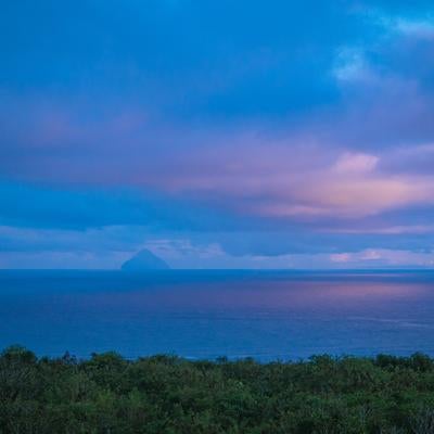 夕焼け色の空と同じ色に染まる海の先にかすむ南硫黄島の写真