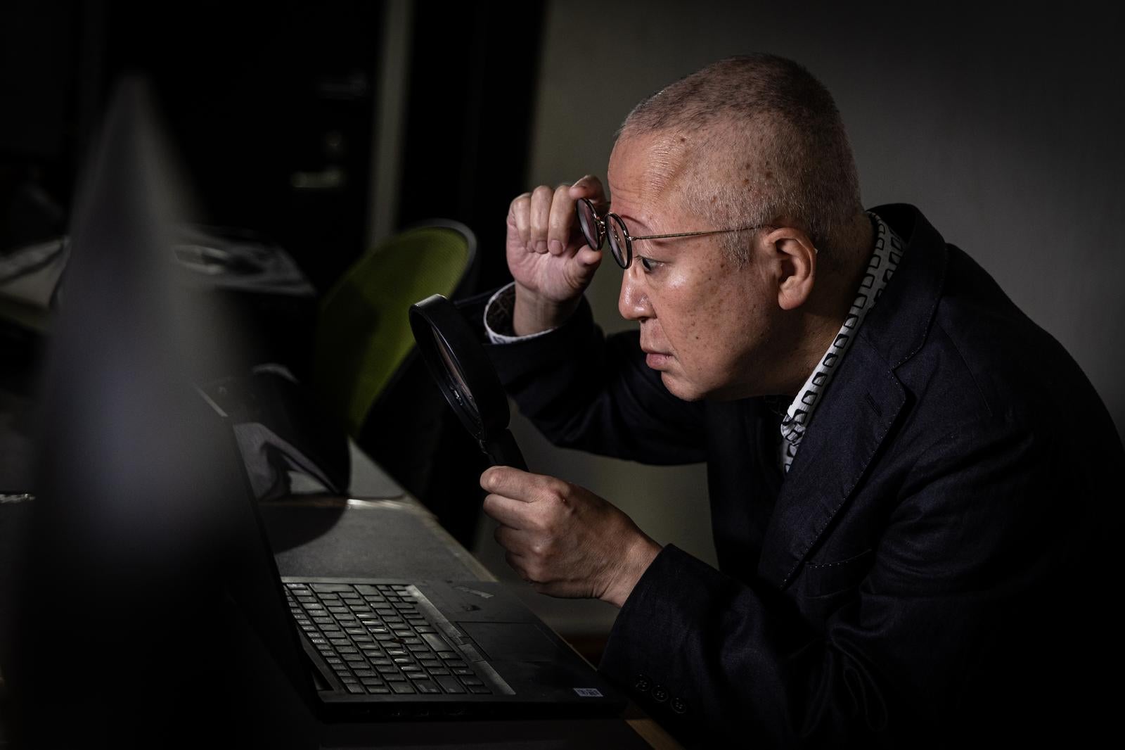 Middle-aged man with short hair removing his glasses and using a magnifying glass to peer at a laptop screen in a dark room