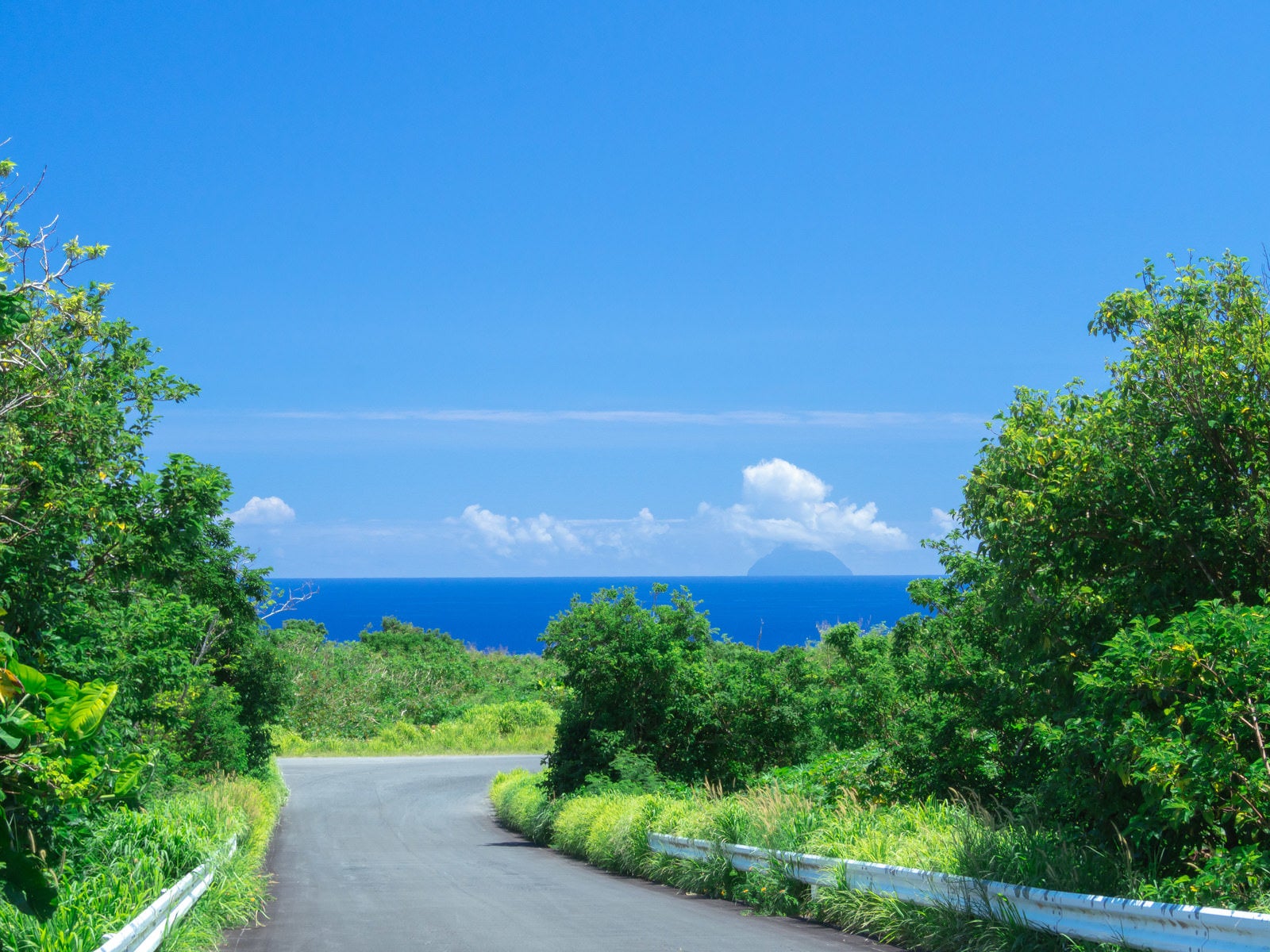 「坂道の先に広がる蒼海と奥にかすんで見える南硫黄島」の写真