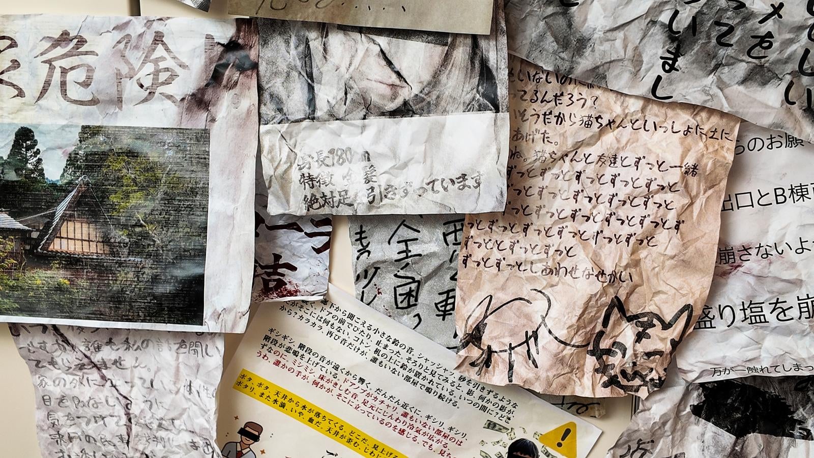 壁一面に貼られたくしゃくしゃの怪文書。危険の文字や手書きの文章、猫の絵などが並ぶ
