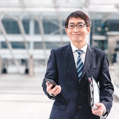 ニコニコしたアラフィフ部長がスマートフォンを片手に携帯電話を持ちながら立つの写真