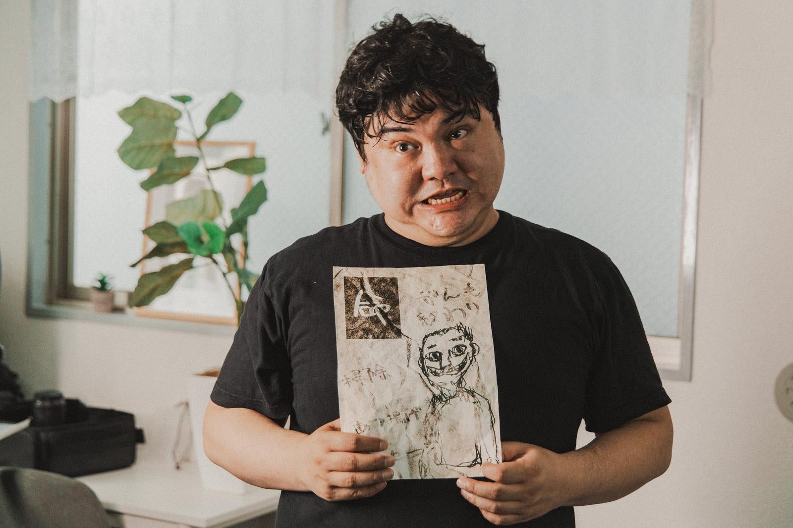 黒Tシャツの男性が奇妙な手書きスケッチを両手で持ち、こわばった表情でカメラを見つめている［モデル：段田隼人］