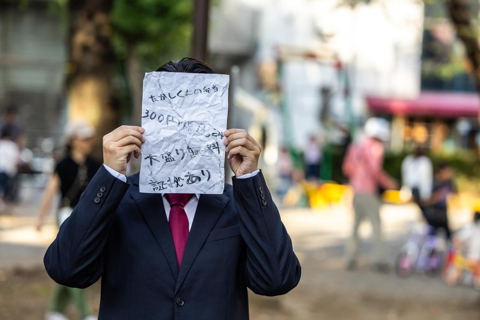 紺スーツ姿の男性が怪文書が書かれた紙で顔を隠す［モデル：大川竜弥］