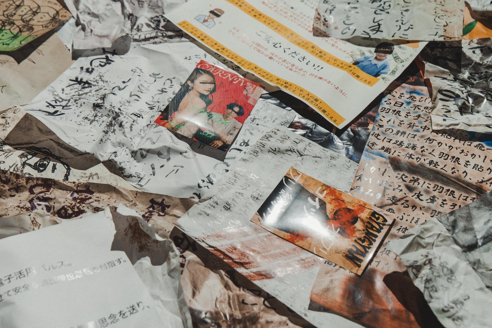黒インクの手書き文字が書かれた紙片が床に散乱し、赤と黄色の印刷物が重なっている