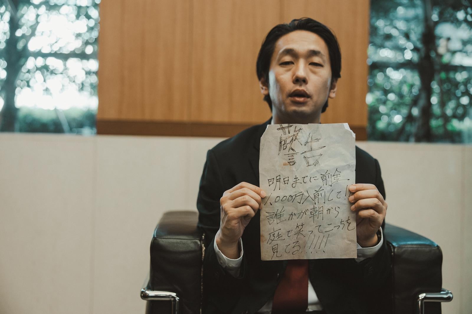 「応接室で怪文書を掲げるスーツ男性」の写真［モデル：大川竜弥］
