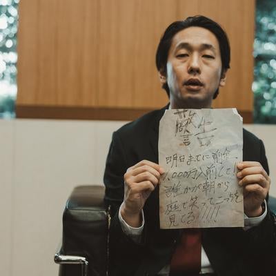 応接室で怪文書を掲げるスーツ男性の写真