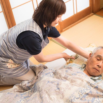 布団の上に横になった老人の枕を直す女性介護士の写真