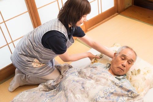 布団の上に横になった老人の枕を直す女性介護士の写真
