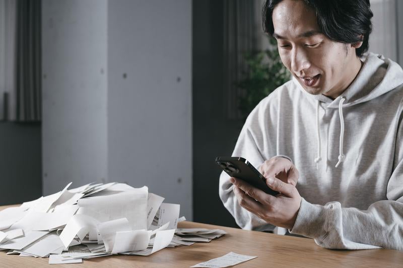 テーブルに散乱した大量の領収書を前にスマートフォンで作業する男性の写真