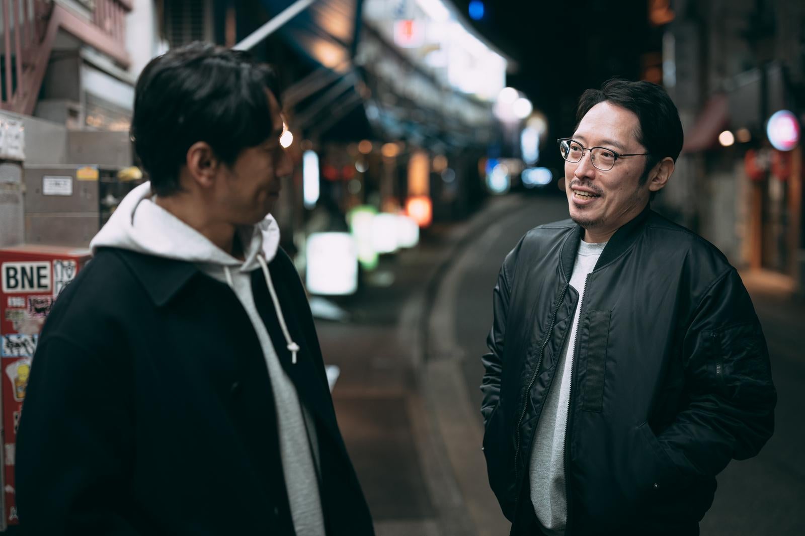 夜の街中で向き合って会話する眼鏡をかけた男性と別の男性［モデル：大川竜弥 ゆうせい］