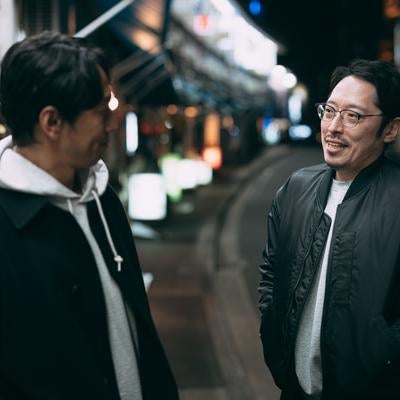 街の喧騒を背に、事件の真相を求めて追跡する二人組の探偵の写真