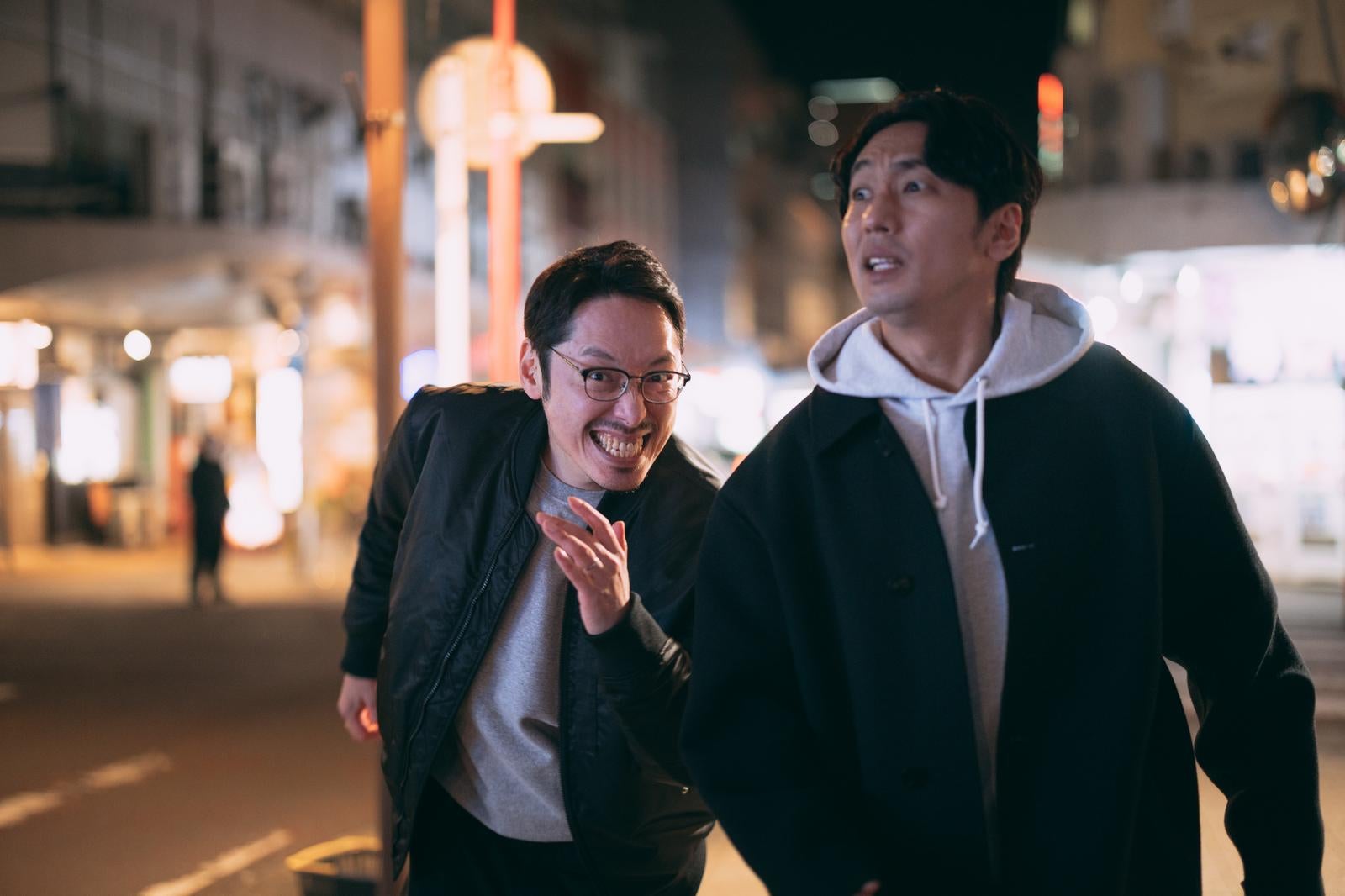 夜の街を歩く眼鏡の男性と、パーカー姿の男性2人が会話している様子［モデル：大川竜弥 ゆうせい］