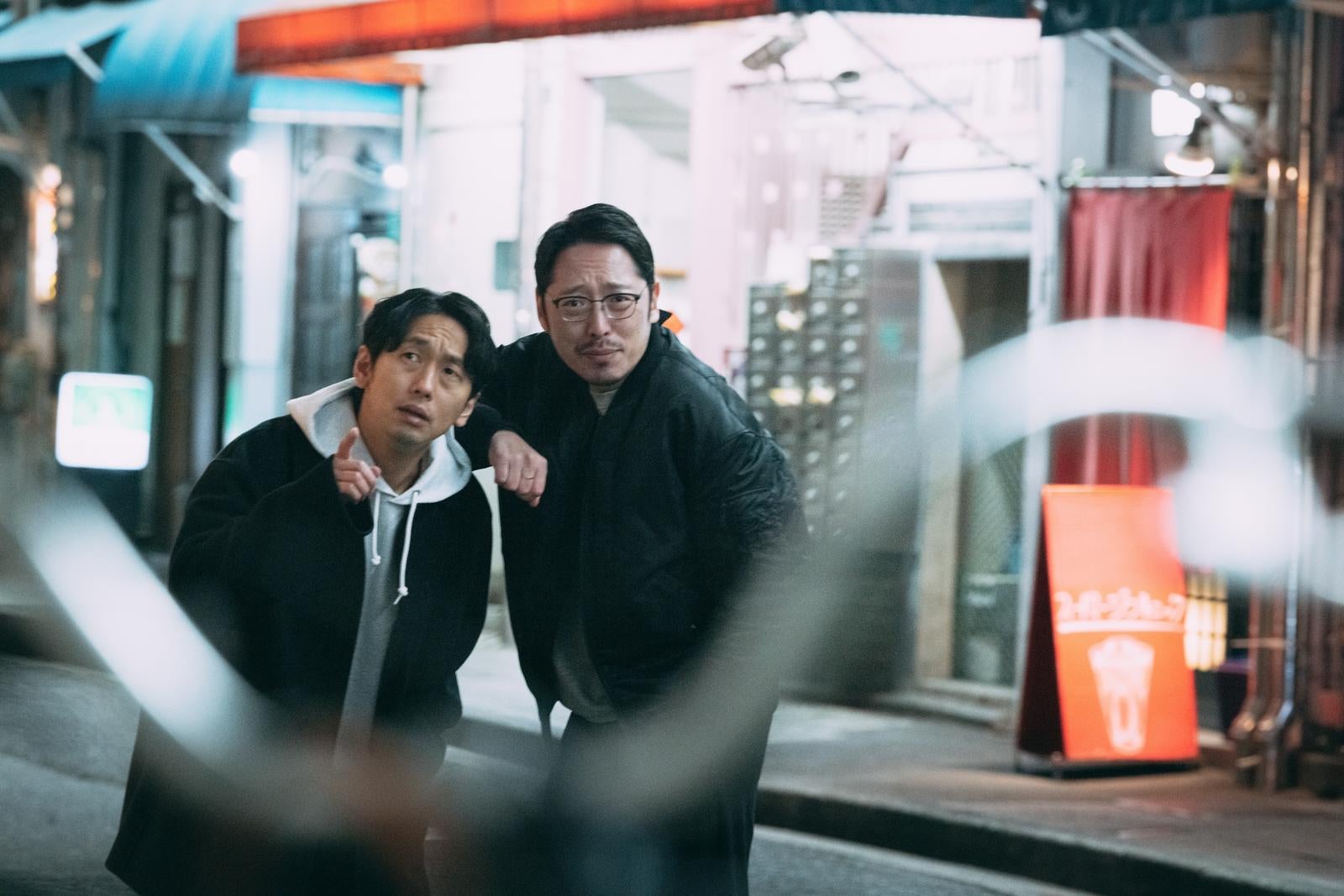 夜間の都市で対象を尾行する黒いジャケット姿の男性二人［モデル：大川竜弥 ゆうせい］