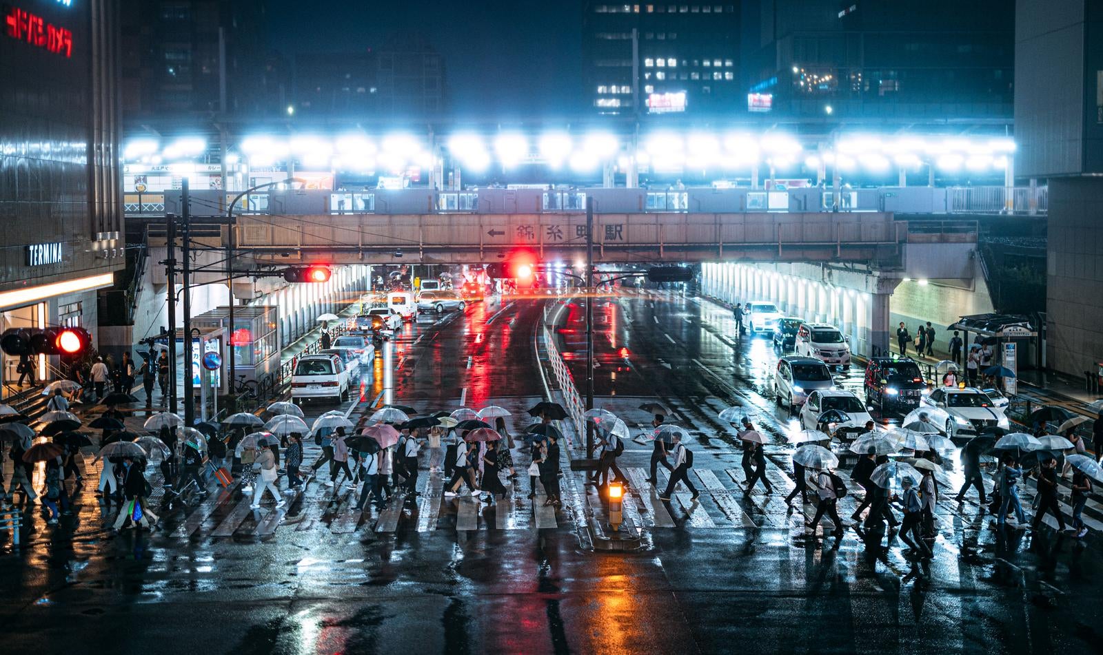 「夜の錦糸町駅前の横断歩道を傘を差して横断する人々」の写真