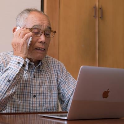 パソコンの使い方を電話で確認しながら操作する高齢者男性の写真