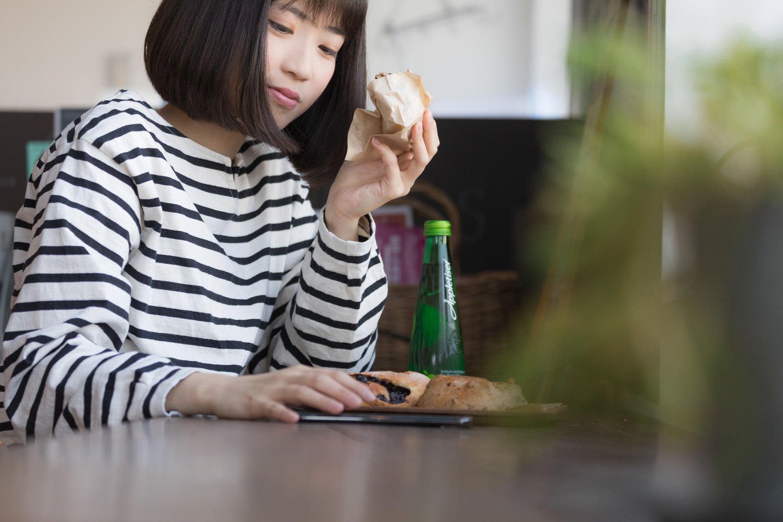 「フードコートでパンを食べながらスマホを見る女性」の写真［モデル：塩田みう］