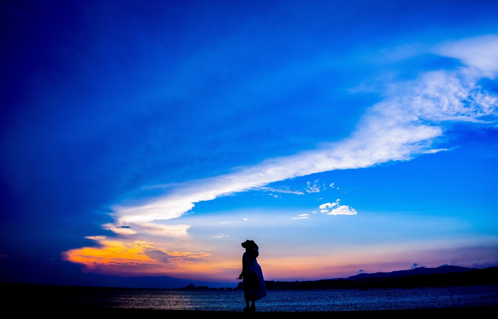 「トワイライトの海岸を歩く帽子をかぶった女性のシルエット」の写真