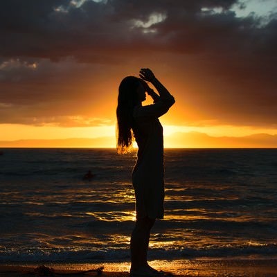 夕日をバックに砂浜で髪をかきあげようとする女性のシルエットの写真