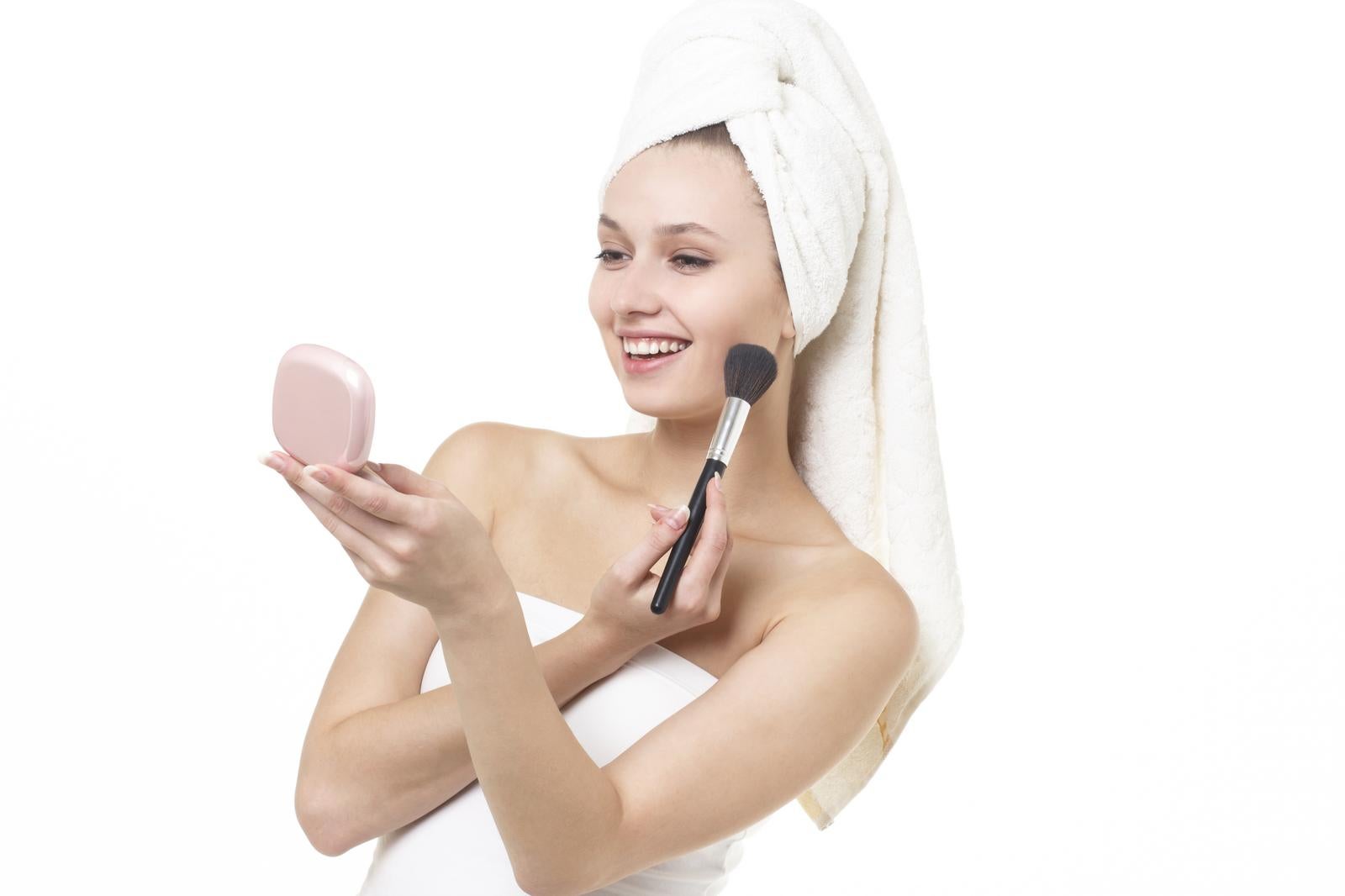 Smiling woman with a bath towel wrapped around her head applying blush with a makeup brush while looking in a compact mirror