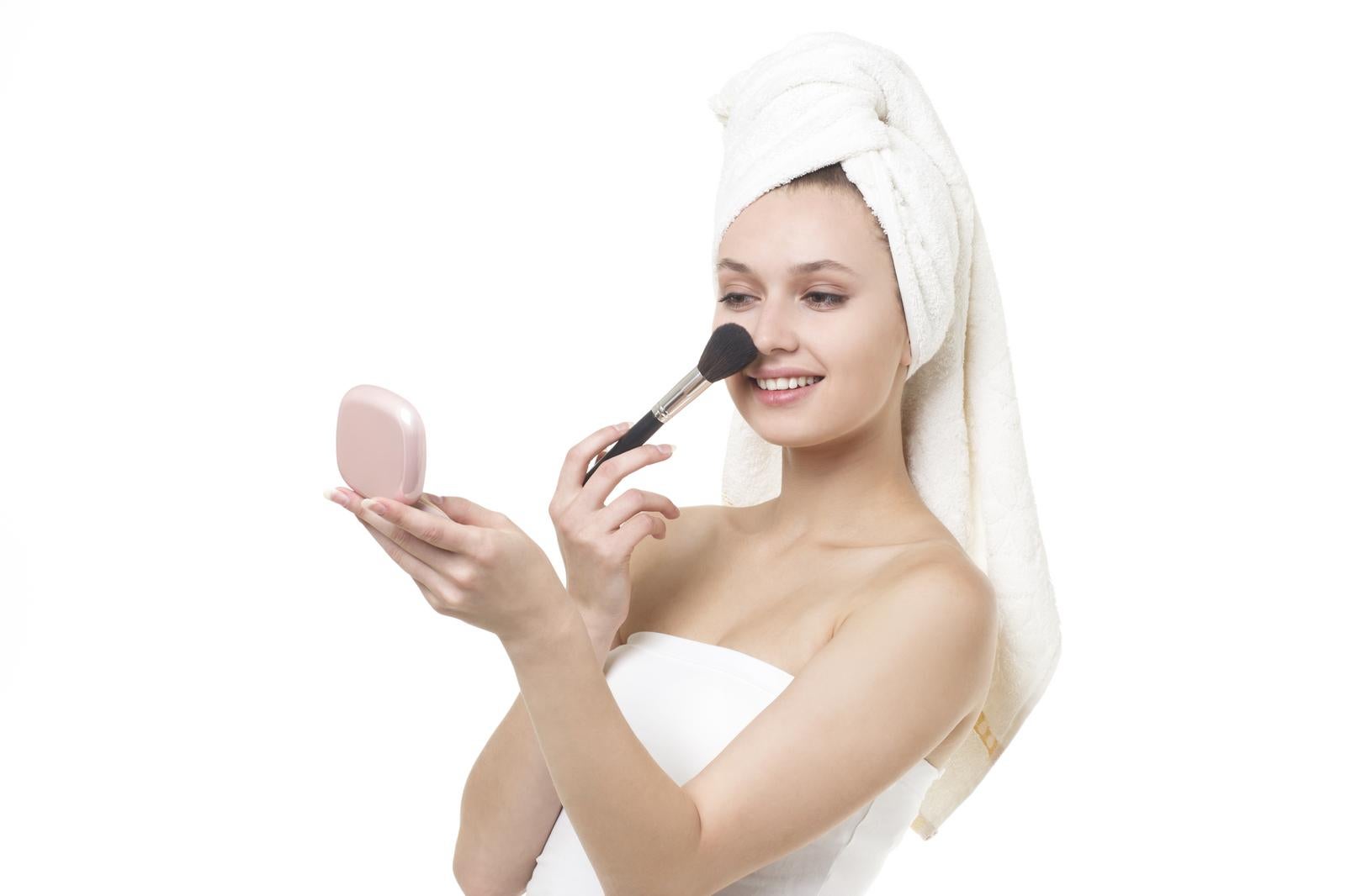 A woman wrapped in a bath towel applying blush to her cheeks with a makeup brush while looking at a compact mirror