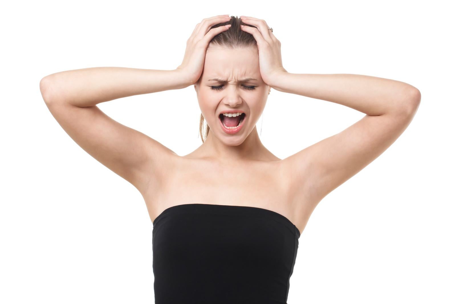 Female model in black bare shoulder top holding head with both hands, screaming with wide-open mouth against white background