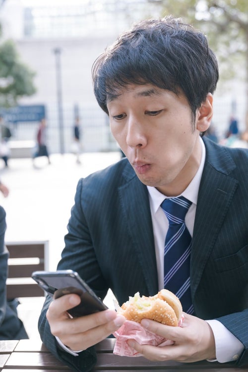 ファーストフードを食べながらスマホをチェックする現代サラリーマンの写真