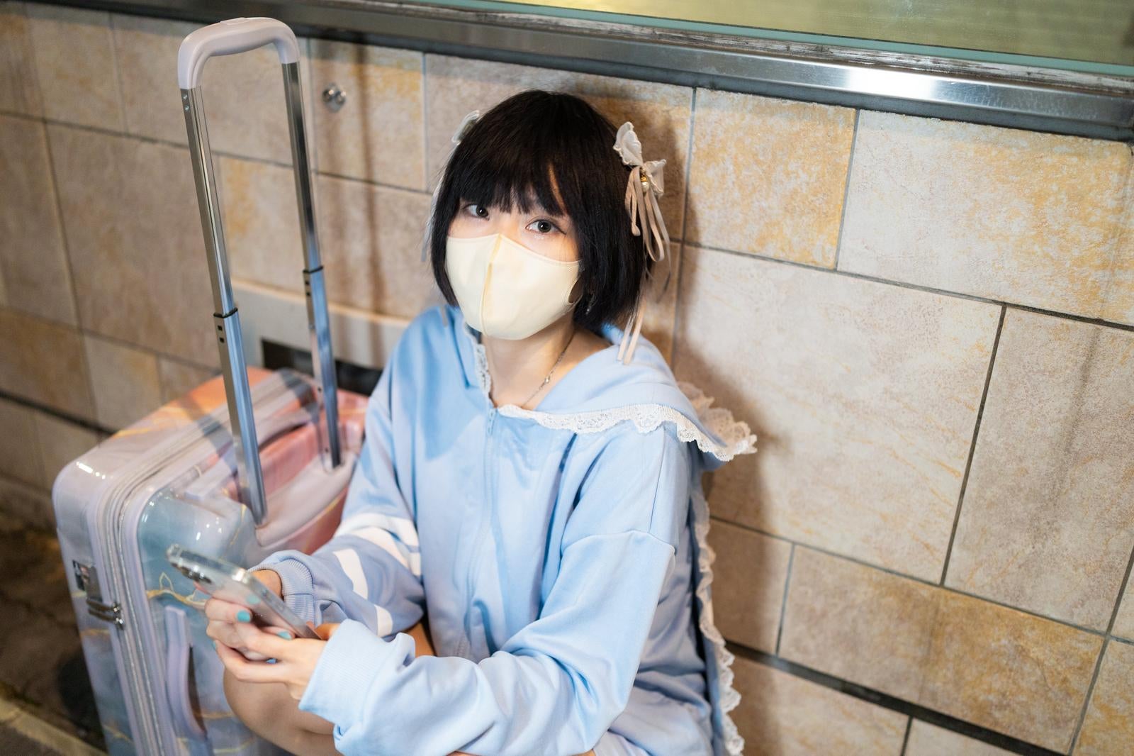 マスク姿でピンクのキャリーケースを持つ黒髪の若い女性が旅行に出発する［モデル：緋真煉］