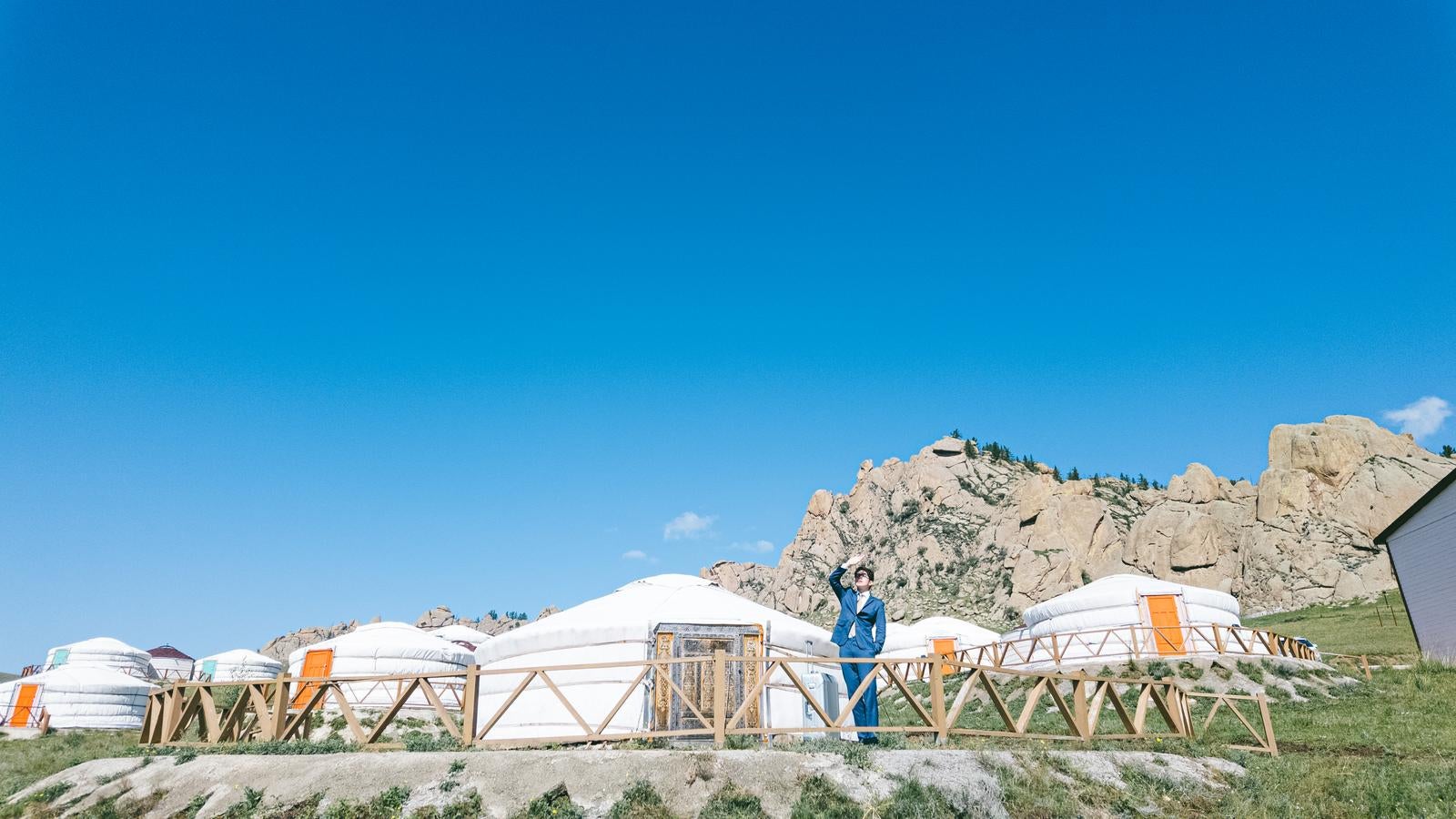 モンゴルの草原に並ぶ白いゲルの前で片手を上げるスーツ姿の男性と背景の岩山［モデル：野崎繁裕］