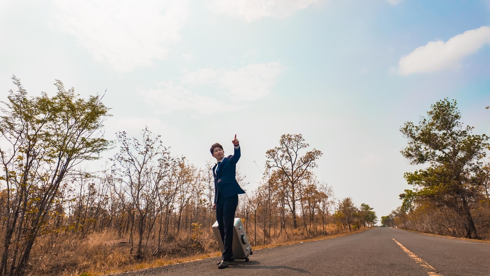 「どこまでも続く一本道でキャリーケースを引くスーツ姿の営業マン」の写真［モデル：野崎繁裕］