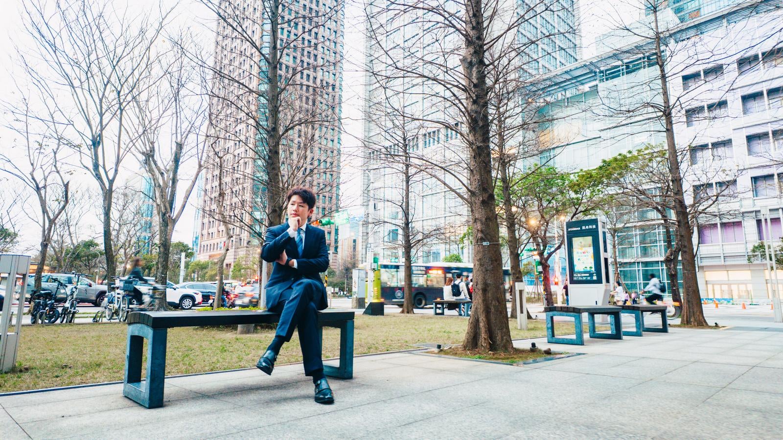 「高層ビルに囲まれたベンチで一息つくビジネスマン」の写真［モデル：野崎繁裕］