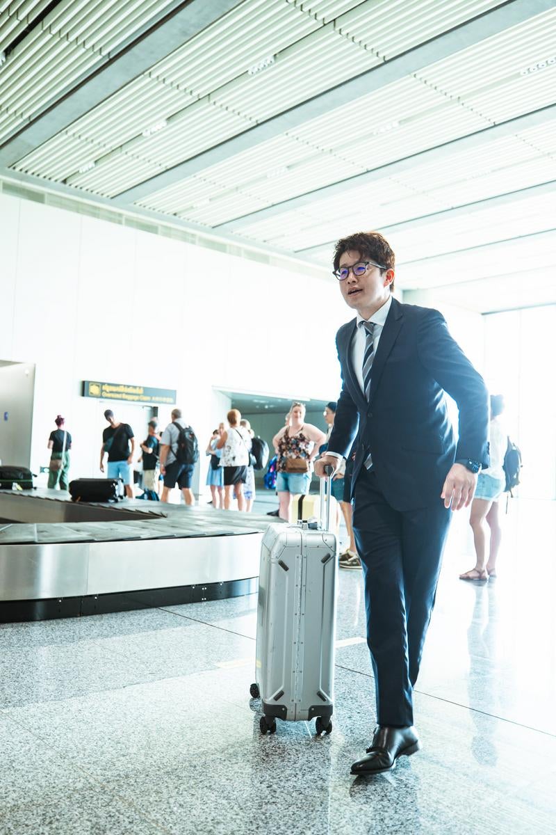 空港ターミナルで白いキャリーバッグを引く紺スーツの眼鏡をかけた男性［モデル：野崎繁裕］