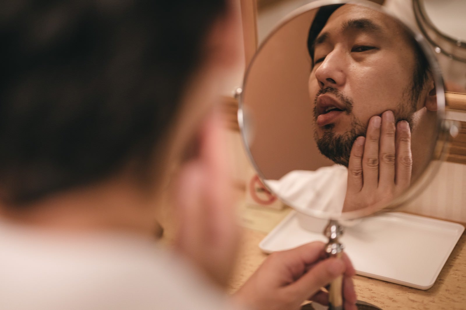 「手鏡で肌のコンディションと伸びた髭をチェックする男性」の写真［モデル：大川竜弥］