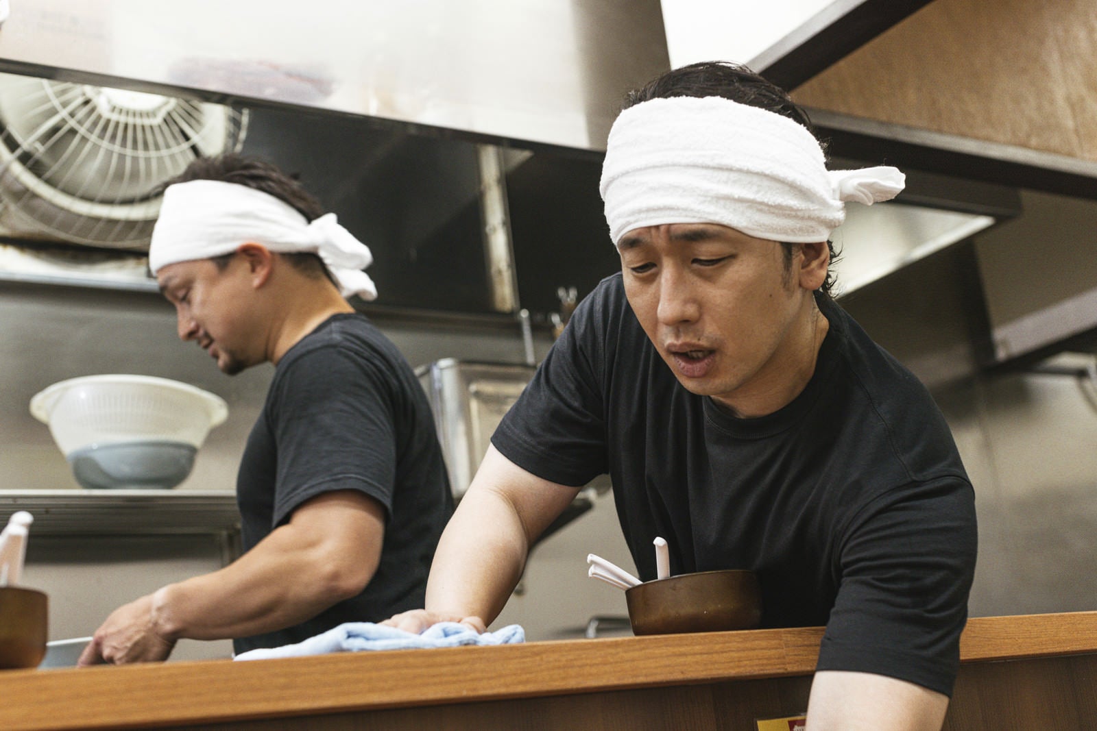 「厨房から体を乗り出してカウンター席を拭くアルバイトの男性」の写真［モデル：大川竜弥 Max_Ezaki］