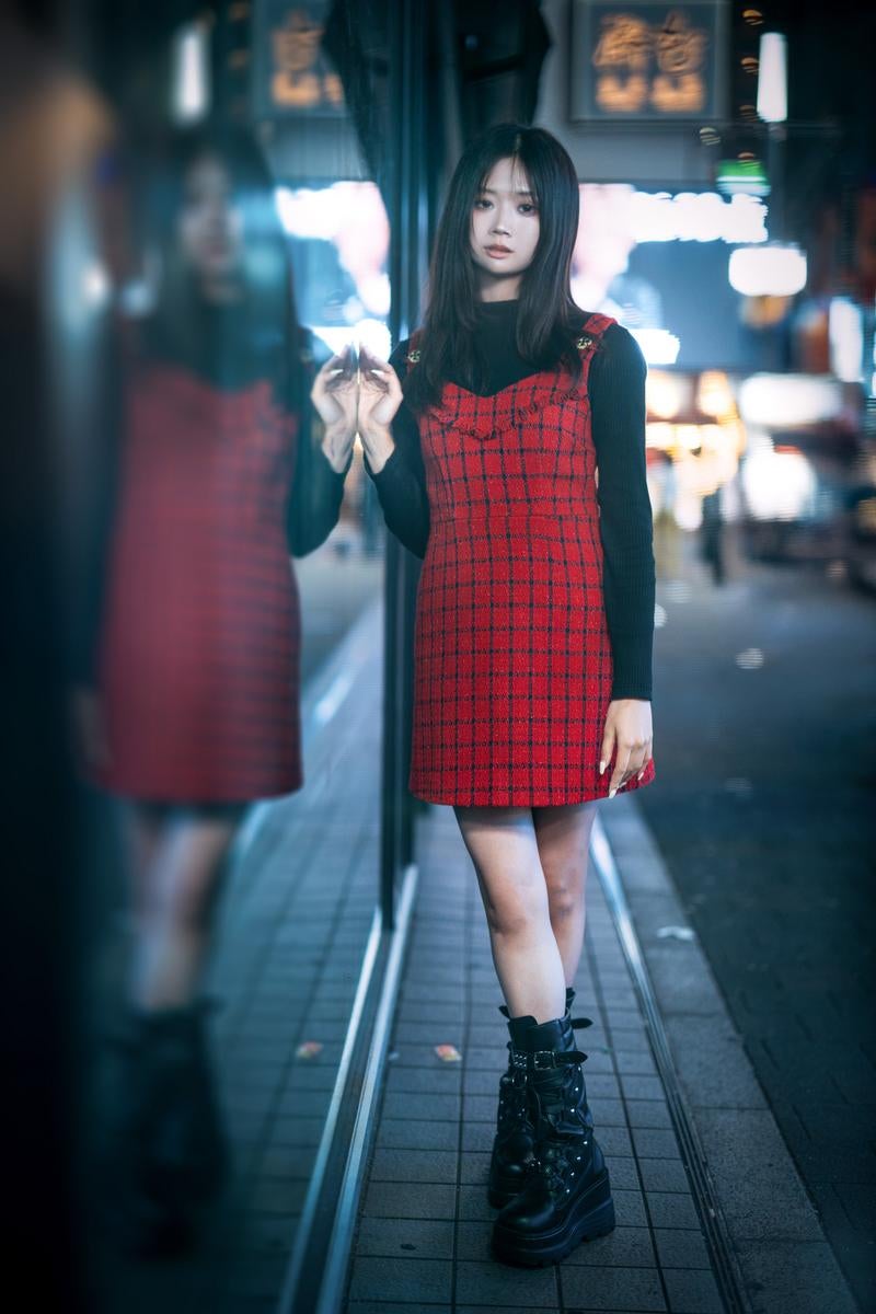 赤いチェック柄のワンピースを着た黒髪の女性が夜間の街角に立ち、背景にはネオン看板と人影がぼけて映っている［モデル：れいか氏］