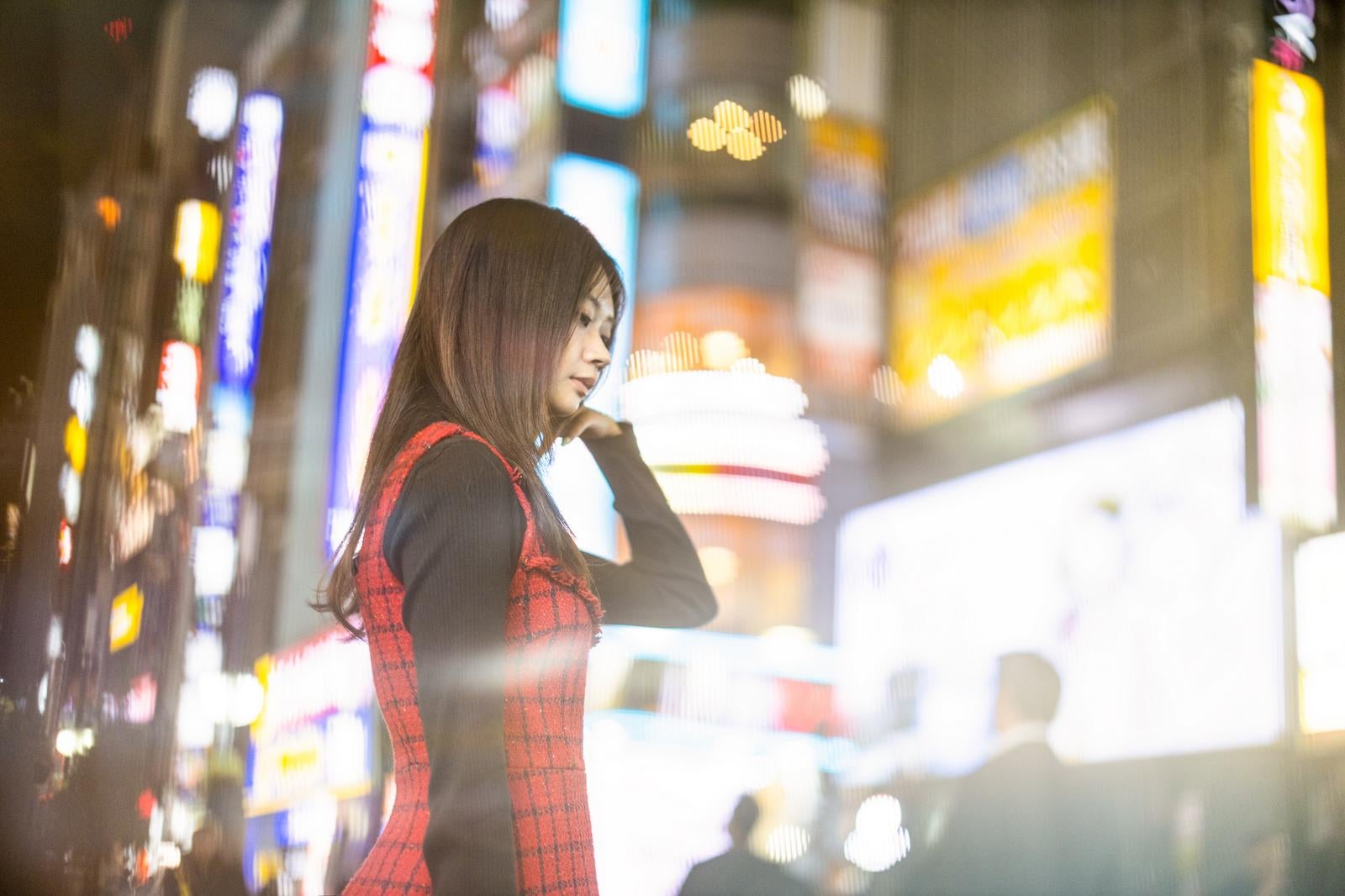 夜の繁華街でネオンをバックに髪に手を添える赤いチェック柄ワンピースの女性のポートレート［モデル：れいかし］