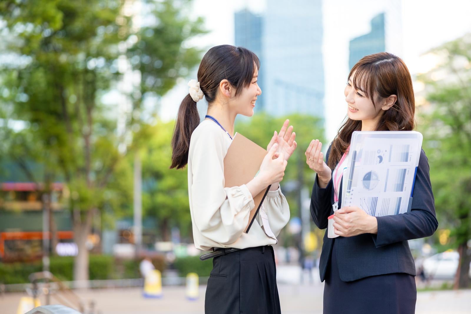 「資料をもって立ち話をする女性社員ふたり」の写真［モデル：SAKI あゆみん］