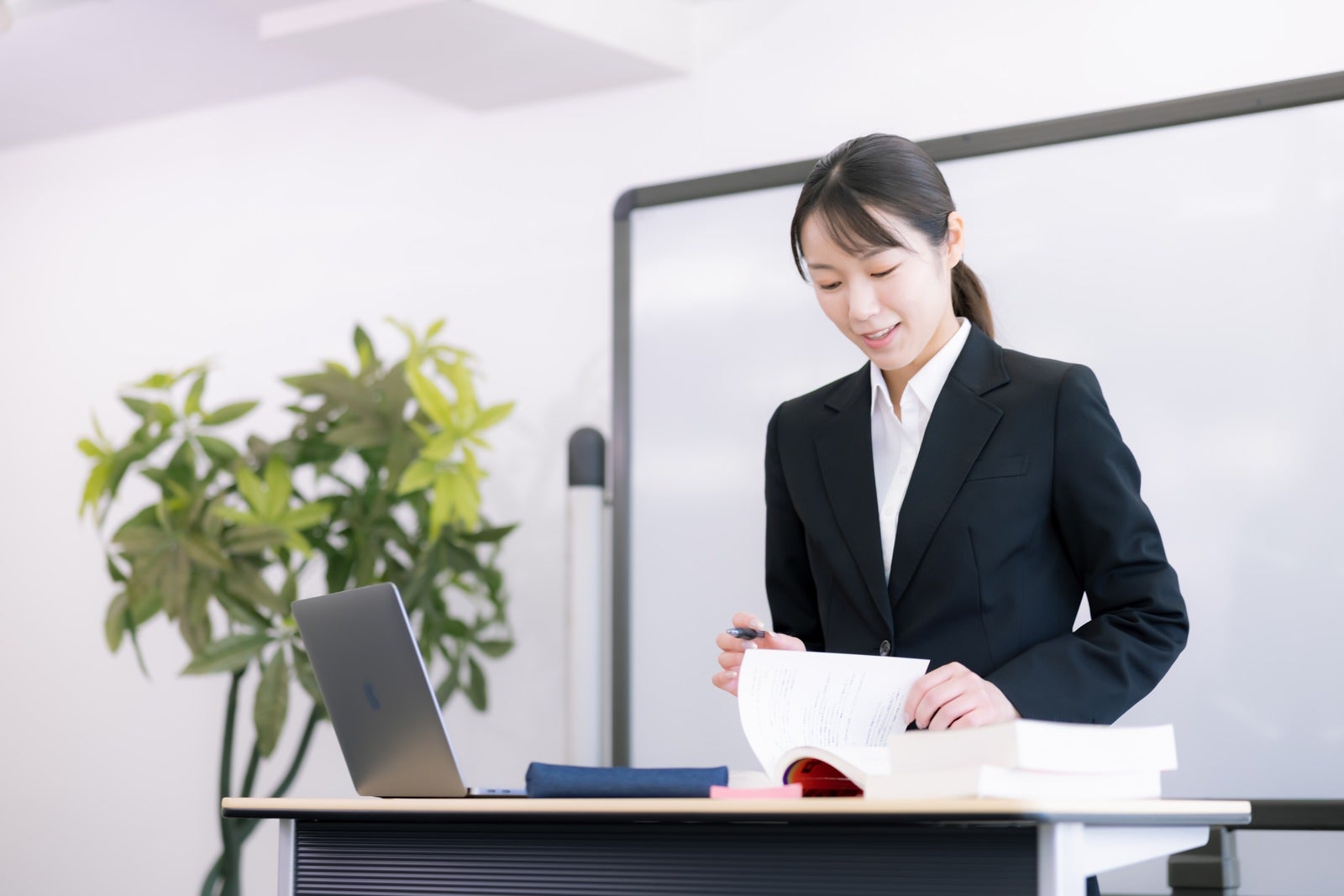 ホワイトボードの前で資料を持ちながら説明するスーツ姿の女性講師［モデル：SAKI］