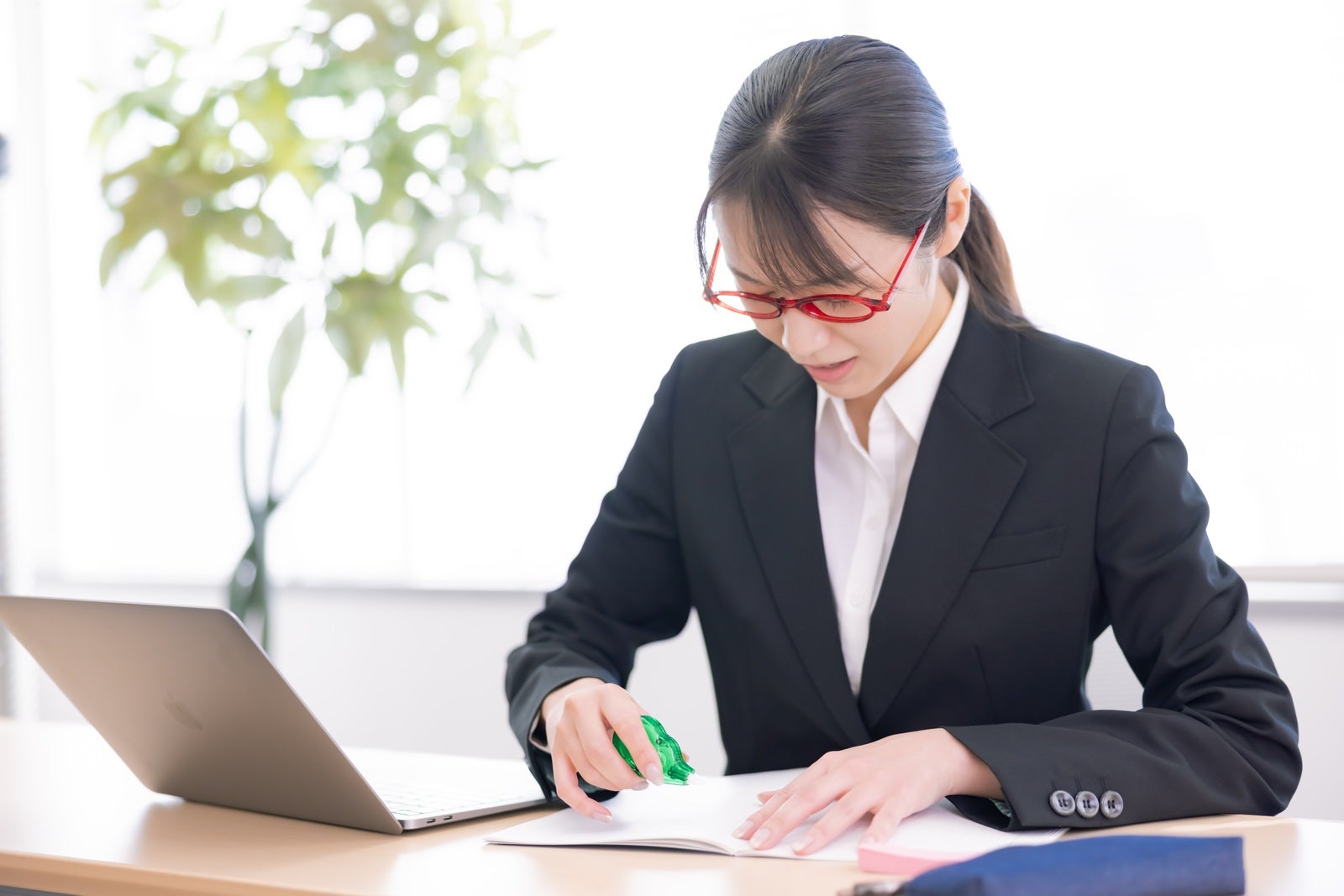 赤いメガネをかけた黒いスーツの女性会社員が修正テープで書類を訂正［モデル：SAKI］