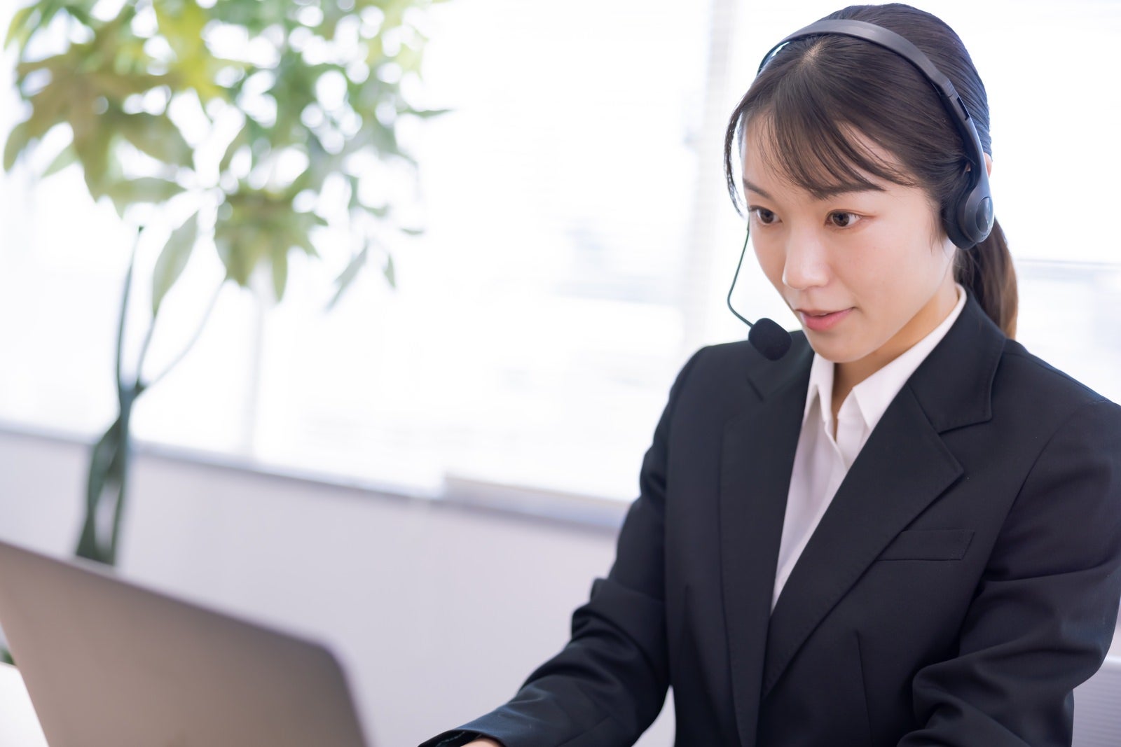 オフィスでヘッドセットを装着しパソコンに向かうスーツ姿のオペレーターの女性［モデル：SAKI］