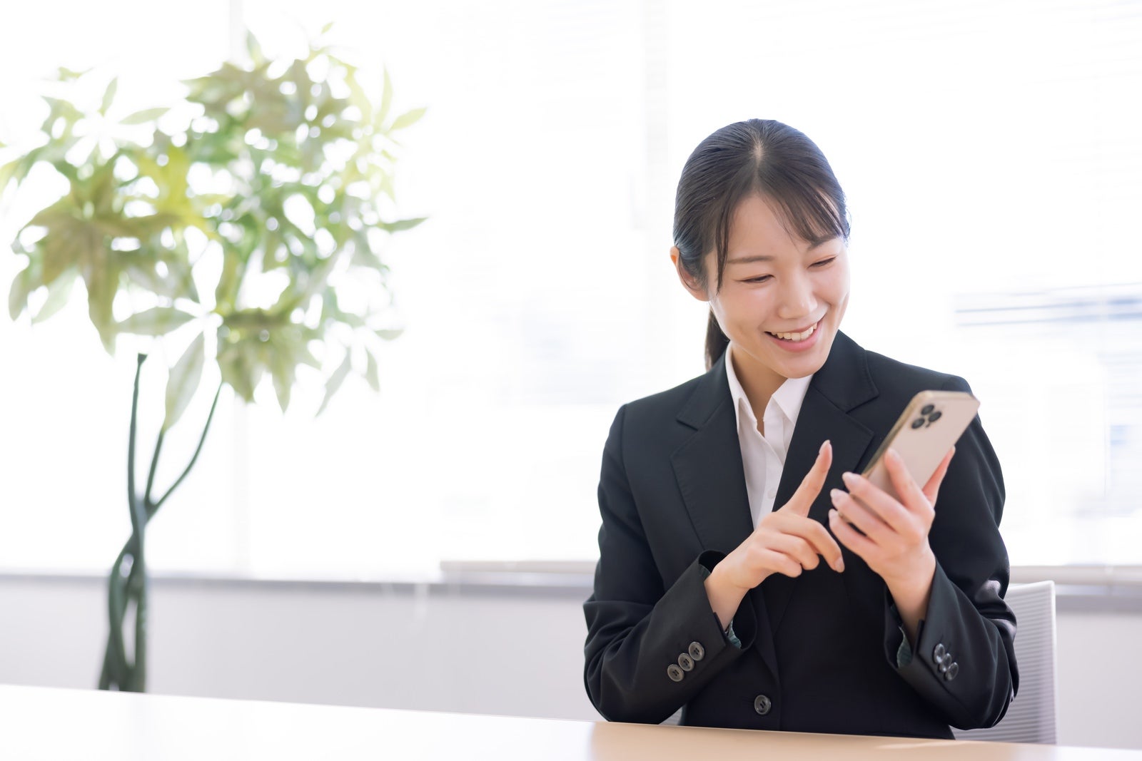 明るいオフィスでスマートフォンを人差し指で操作しながら笑顔を見せるスーツ姿の女性［モデル：SAKI］