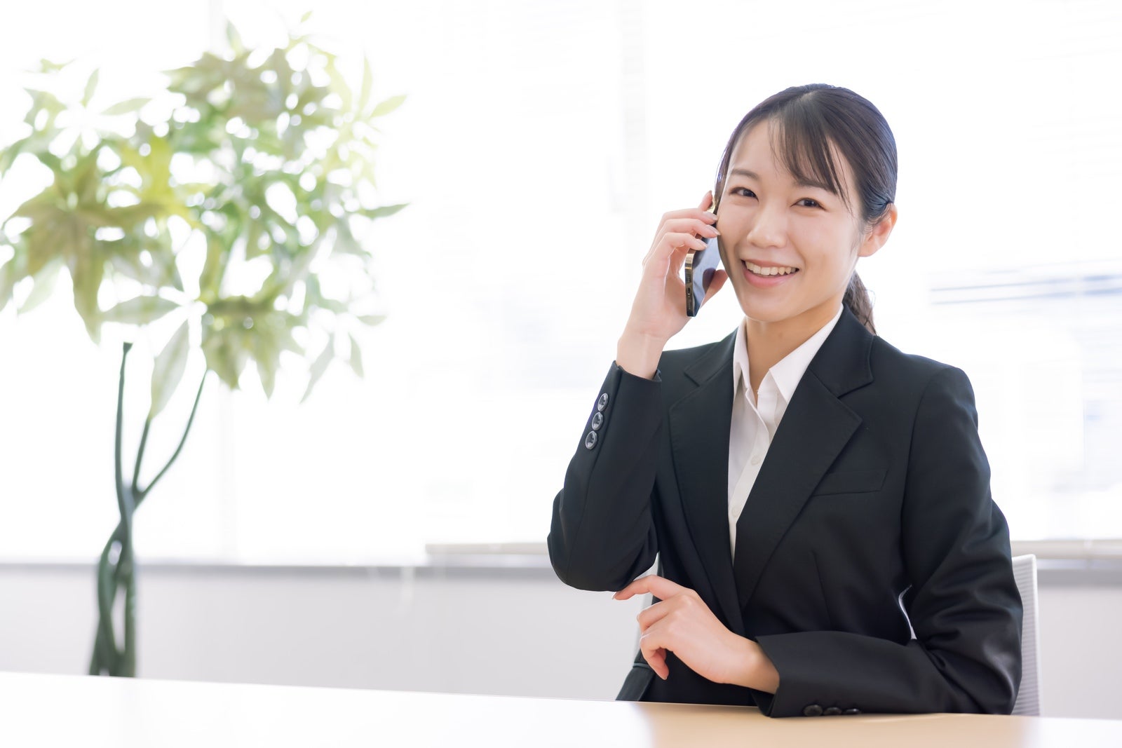 明るいオフィスでスマートフォンを使って笑顔で通話するスーツ姿の女性［モデル：SAKI］