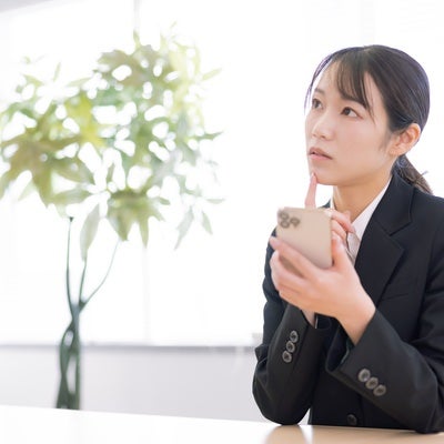 スマホを見て困惑する黒いスーツの女性オフィスワーカーの写真