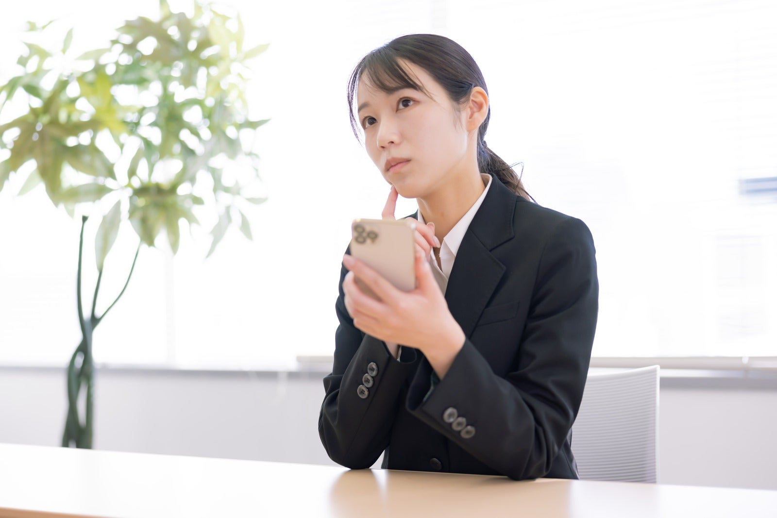 オフィスで困った表情でスマホを見つめる黒いスーツ姿の女性［モデル：SAKI］