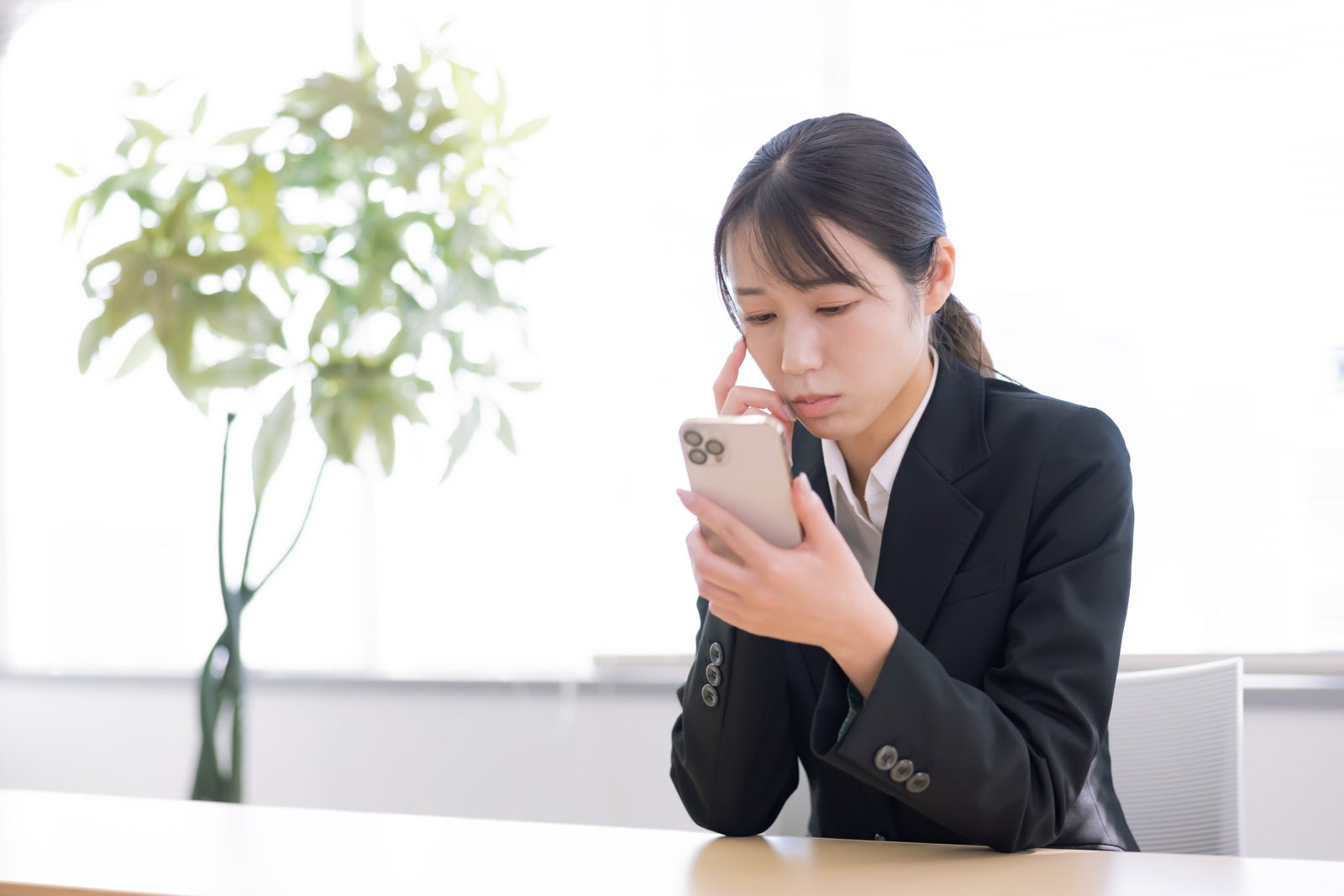 黒いスーツの女性がスマホを持ち、口元に指を当てて困った表情で画面を見ている［モデル：SAKI］